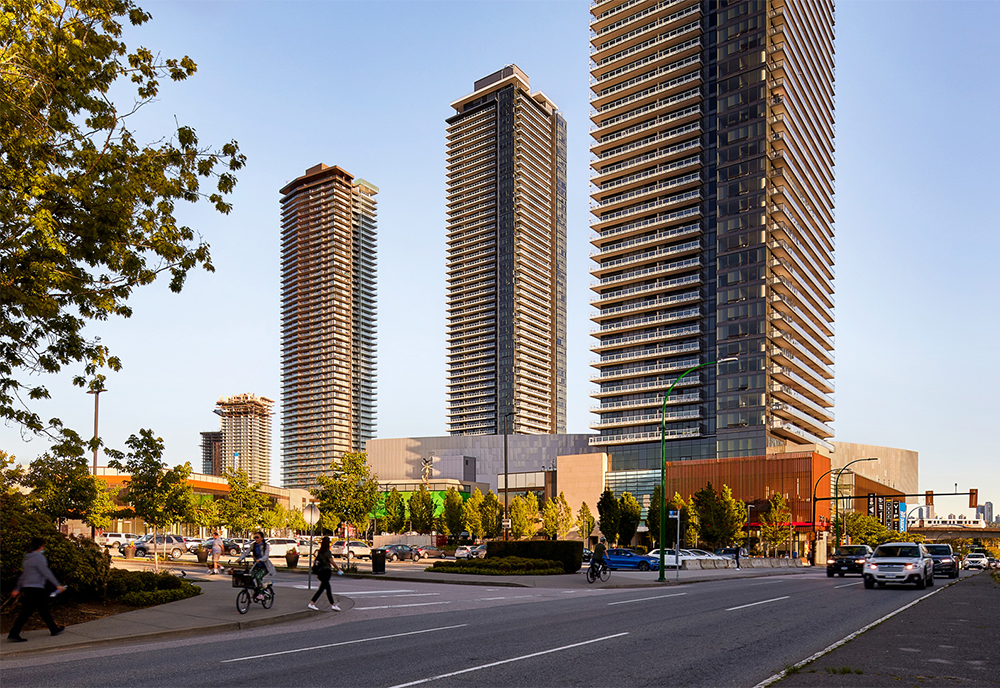 The three Amazing Brentwood towers showing increasing in size from left to right against an evening sky with people walking on the sidewalk in the foreground.